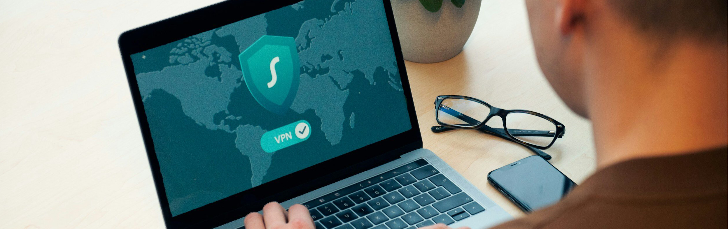 Man typing on computer requiring a vpn with phone and plant in background